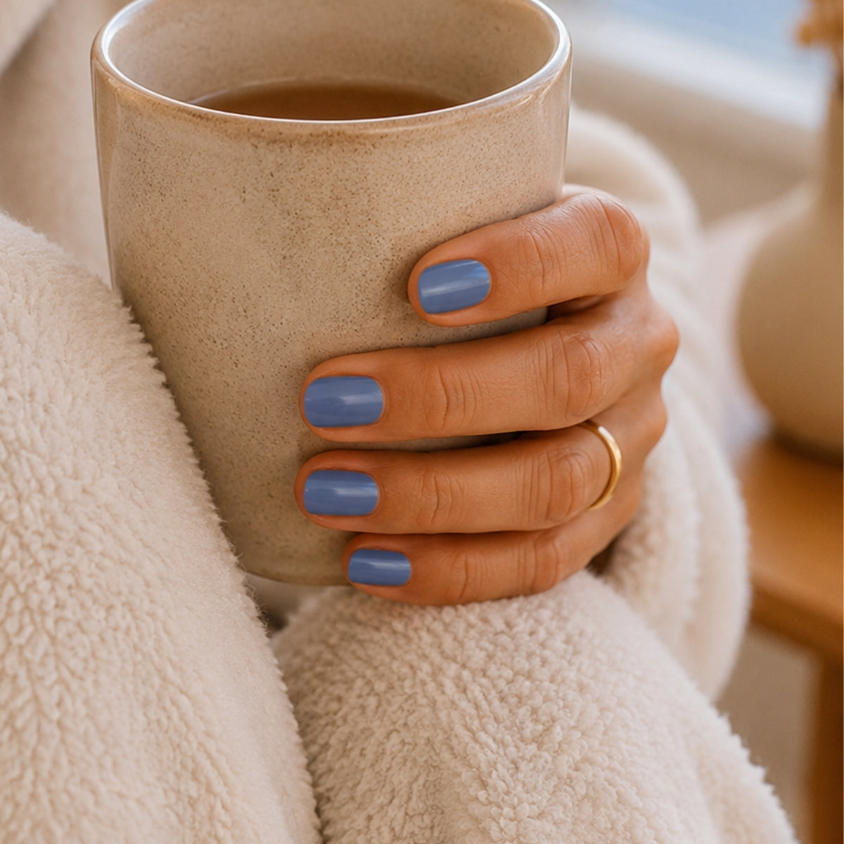 Pastellblaue Nägel im Fokus, die eine Teetasse in der Hand halten. Im Hintergrund sitze sieht man Beine und ein Fenster.